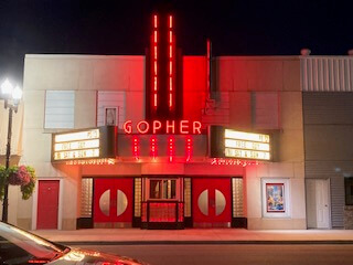Front view of the Gopher Theatre at night, illuminated with bright red neon lights and a glowing marquee, with a car passing in the foreground on the street.