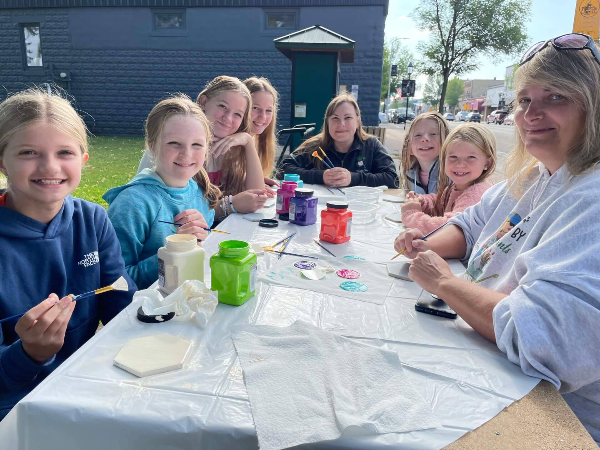 Seven girls and one adult woman around a table outdoors. The tile has painting supplies on it. Everyone is smiling.