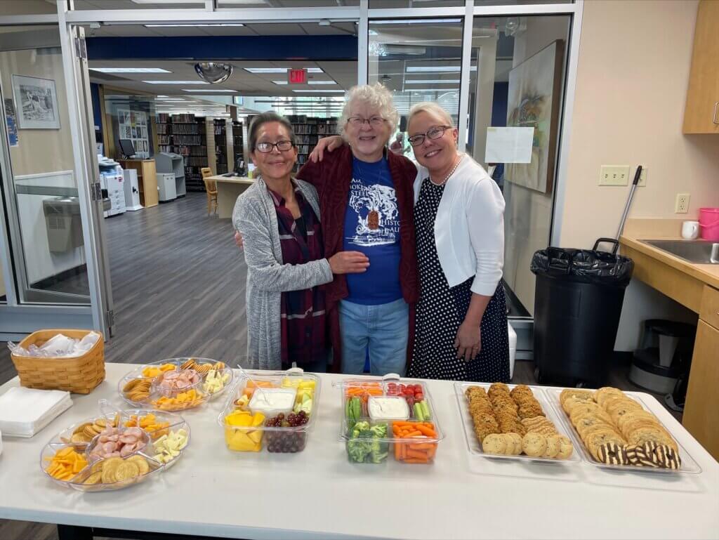 Three ladies standing side by side smiling behind a display of food.