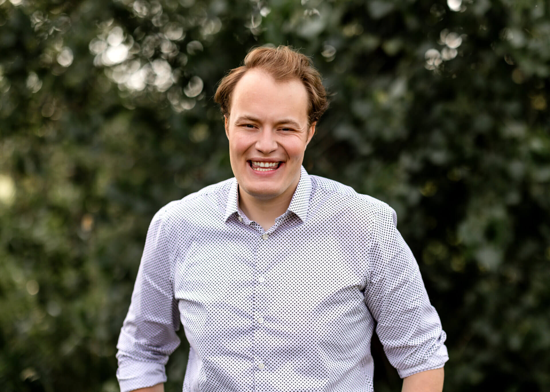 Brogan is a young man with light brown hair. He is wearing a light color button up shirt and standing in front of green trees. He is smiling.