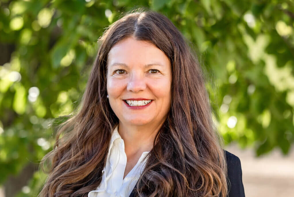 Camille is smiling with long brown hair. She is wearing a white shirt and black blazer. She is standing in front of a green tree.