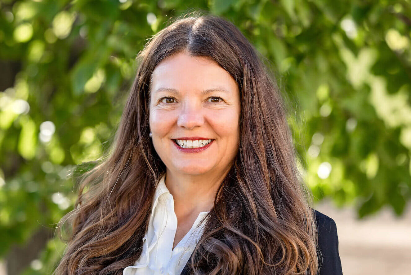 Camille is smiling with long brown hair. She is wearing a white shirt and black blazer. She is standing in front of a green tree.