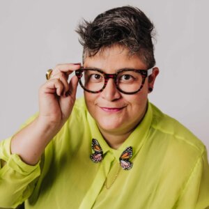 Kandace is wearing a bright yellow-green button-up shirt. They have short dark hair and glasses.