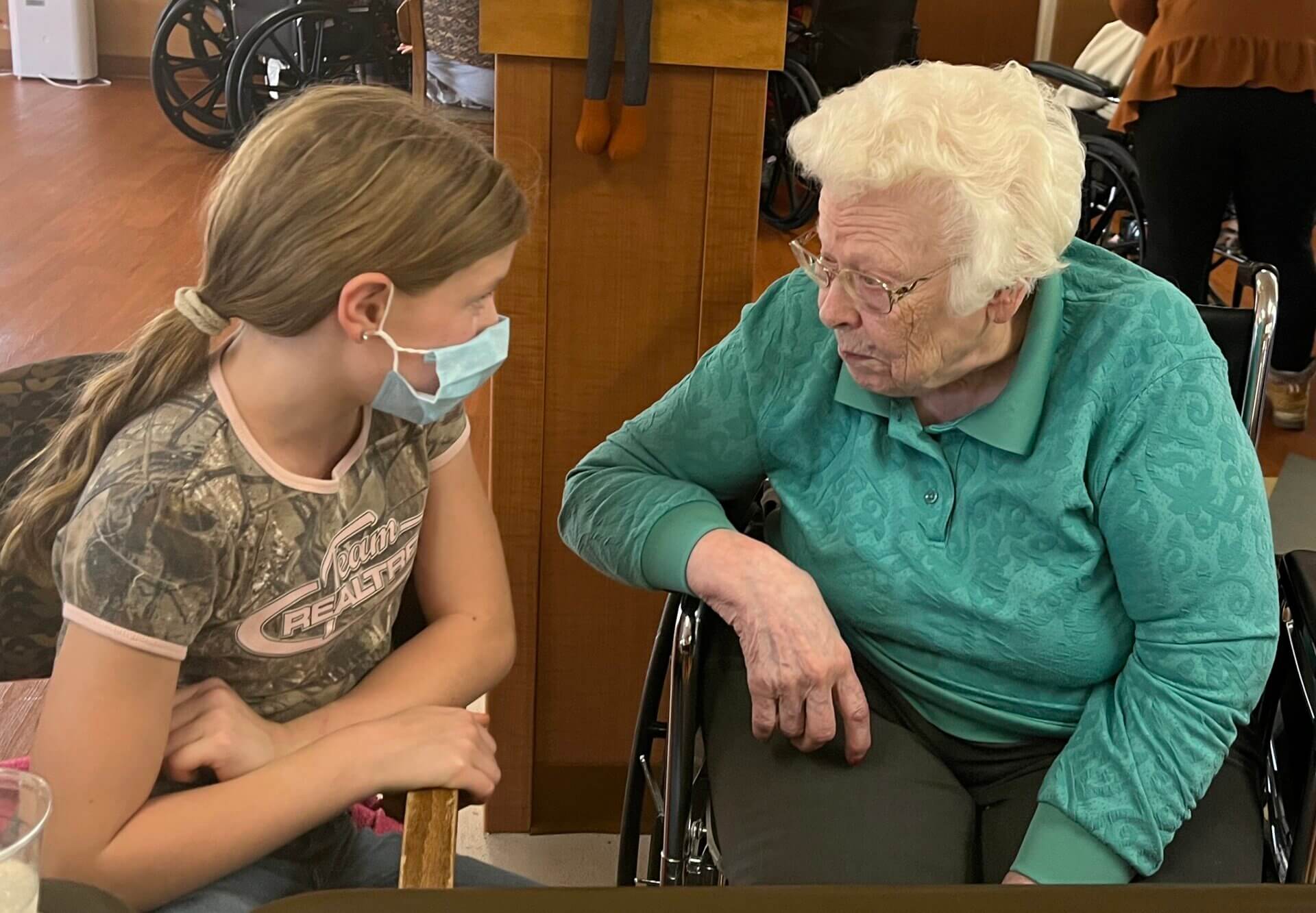 A girl wearing a mask talking closely with an older lady in a wheelchair.