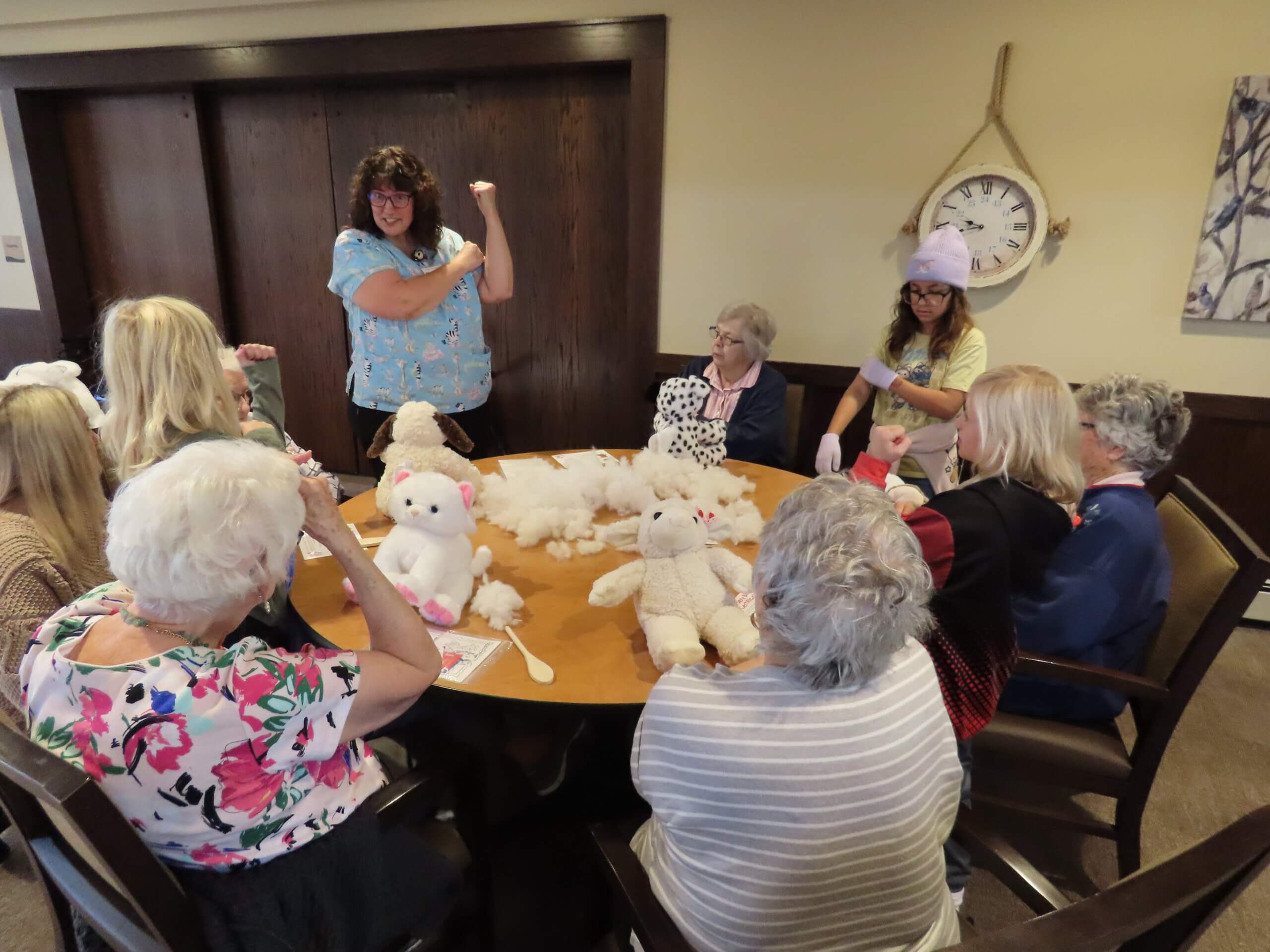 Older adult women and young girls sit around a table that has stuffed animals on it.