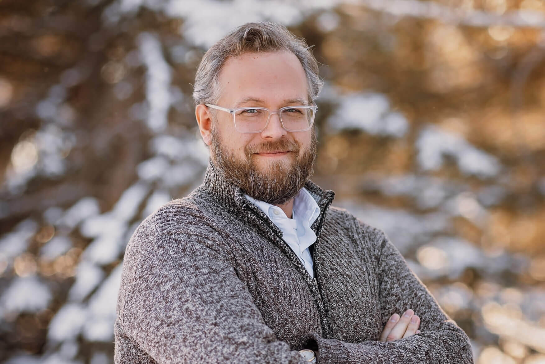 Phil is standing outside on a winter day. He is wearing a neutral color sweater and has his arms crossed in front of his chest. Phil is wearing glasses, has a beard, and is smiling.
