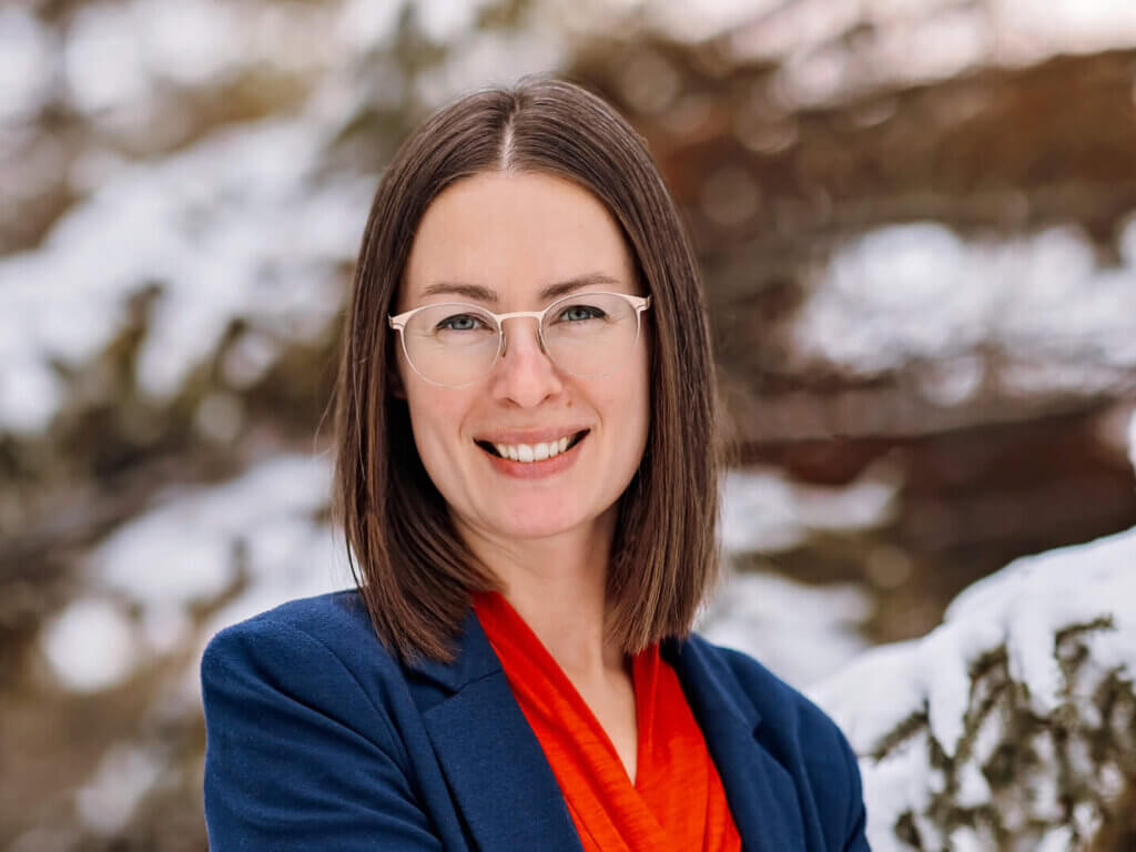 Cedar is wearing glasses and smiling. She has shoulder length straight brown hair. She is wearing a red shirt and blue blazer. She is standing outside on a sunny day in the winter.