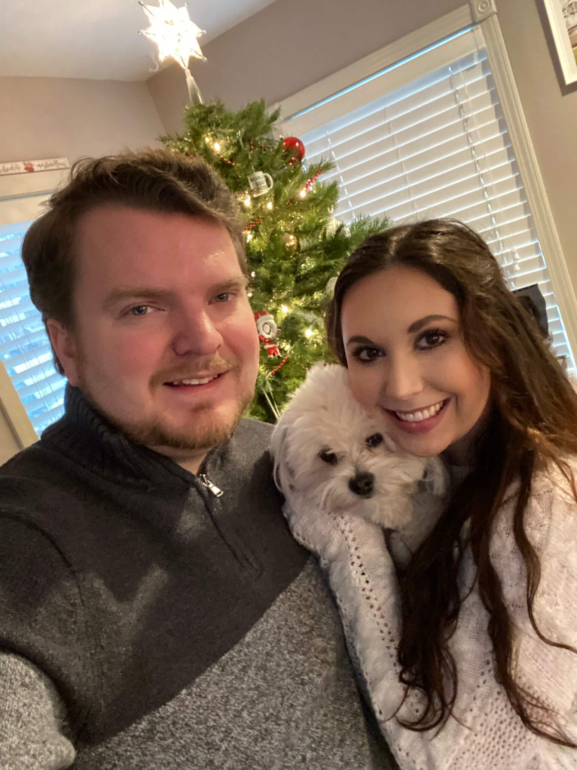 Communications Intern Stella poses with her small white dog and her husband. She has long brown hair, is wearing a white sweater, and is smiling.