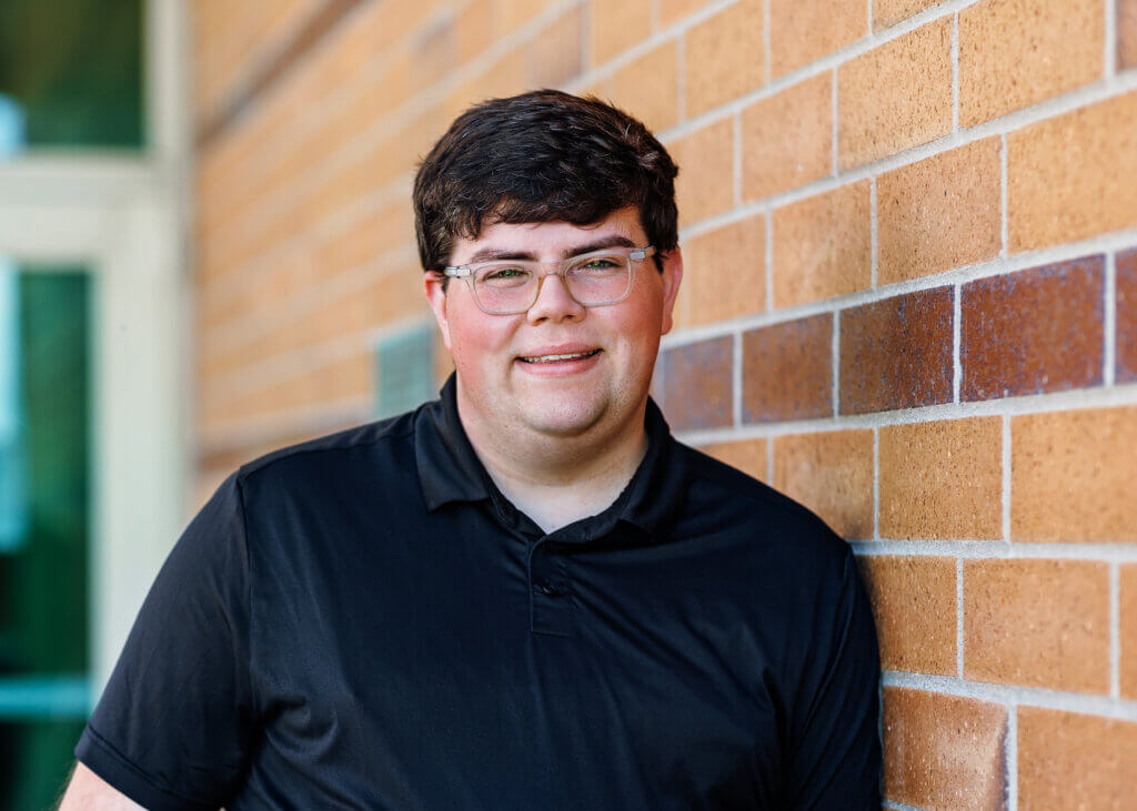 Griffin is wearing a black polo shirt and clear glasses. They have short brown hair. Griffin is smiling and leaning against a red brick wall.