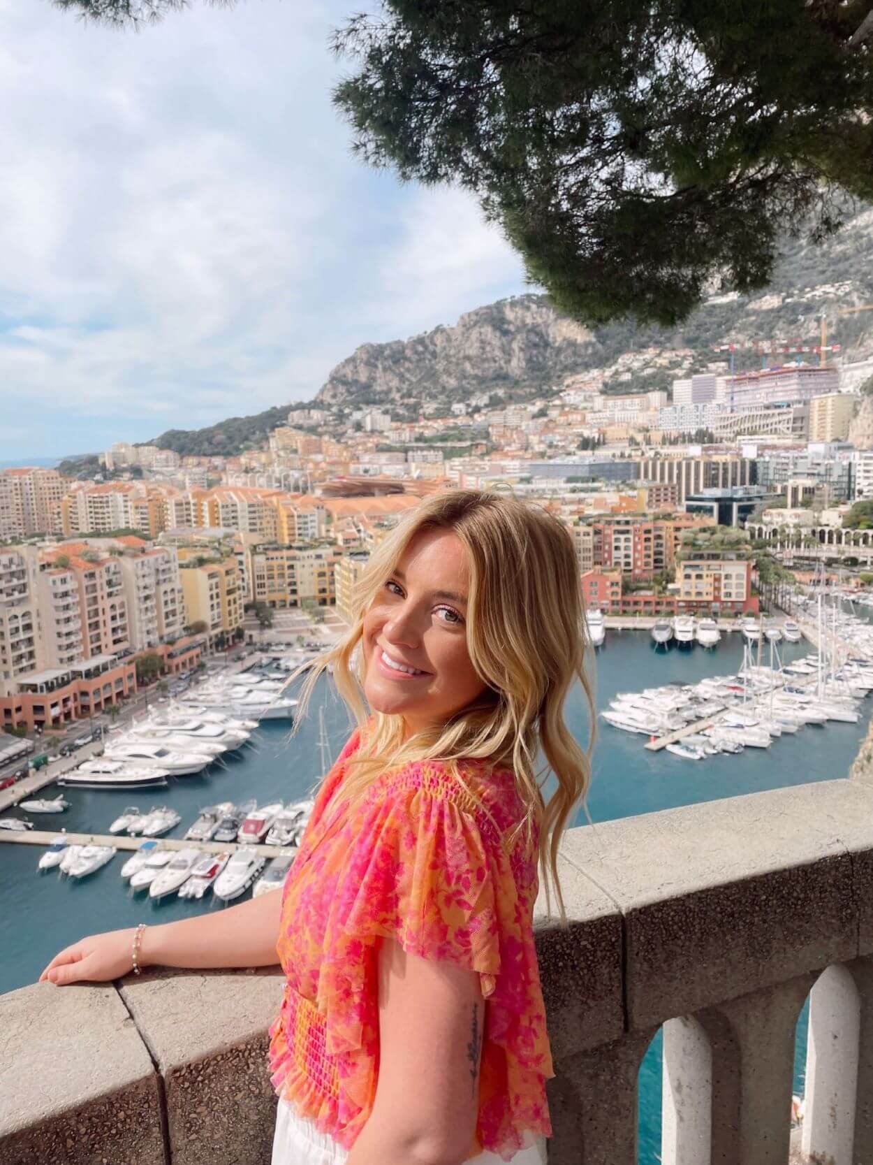Communications Intern Greta Hentges poses in front of a city scape in Italy. Greta is blonde, wearing a pink and orange floral shirt, and is smiling at the camera.
