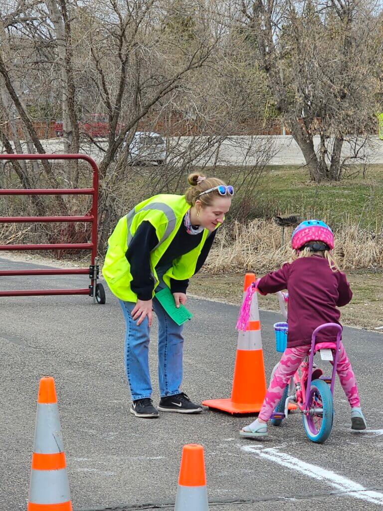Mallory Jarvi is wearing a bright green traffic vest and helping a child wearing pink pants, shirt, and helmet on a pink bike.