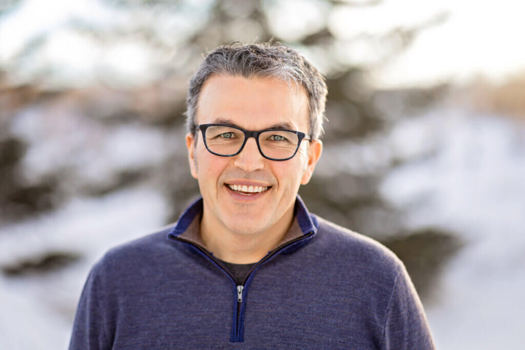 Ryan Rooney stands outside on a snowy day. He is smiling. He is wearing a blue quarter-zip sweater and glasses. He has short salt-and-pepper hair.