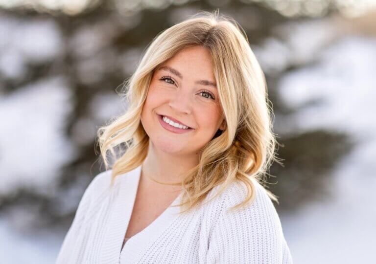 Greta is wearing a white sweater. She has long blonde hair and is standing out in the snow smiling.