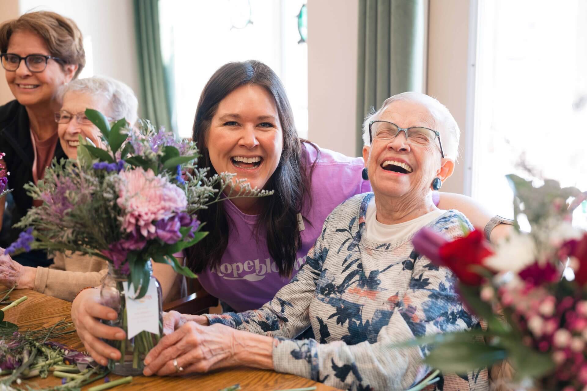 Two women are laughing around a table with flowers.