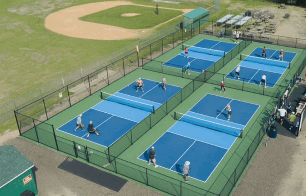 This image is taken from above. It shows four blue and green pickleball courts surrounded by a black chain link fence. There are people playing pickleball on the courts.