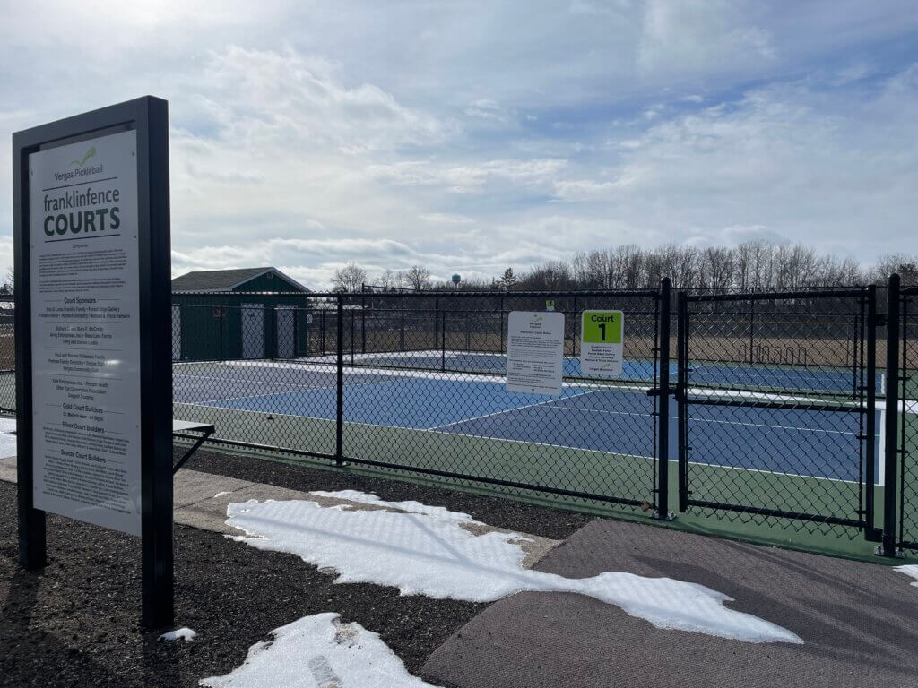 There are four blue pickleball courts. A tall black chain link fence surrounds the courts. A tall sign explains that the courts are Vergas' pickleball courts.