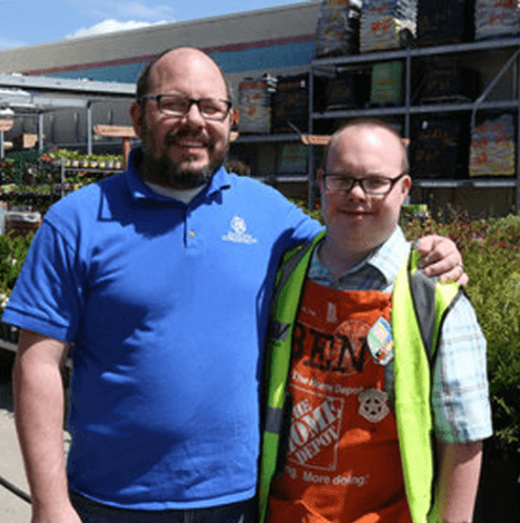 Two men are standing and smiling at the camera. One is wearing a bright blue shirt, and the other is wearing an orange Home Depot apron and a yellow safety vest.