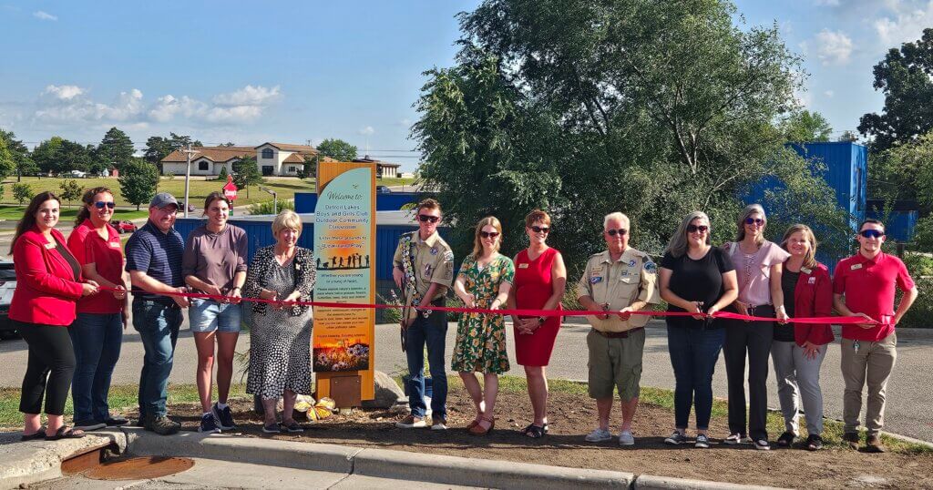 Pollinator Garden Ribbon cutting at Boys & Girls Club of Detroit Lakes. 13 people stand outside holding a long red ribbon and scissors.