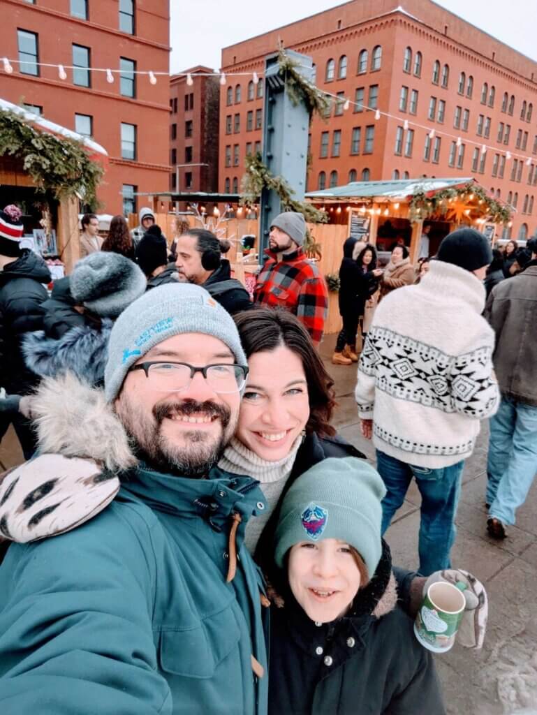 Jenna, Pete, and Max stand outside in a city in the winter. Pete and Max are wearing hats.